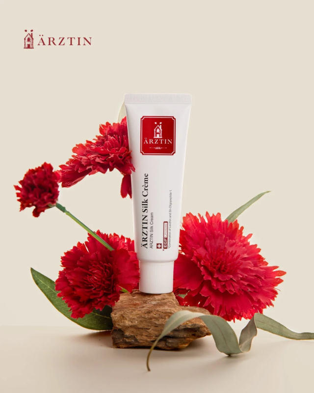 La crema de seda de &Auml;RZTIN junto a una flor de clavel, mostrando su envase elegante y su conexi&oacute;n con ingredientes naturales.