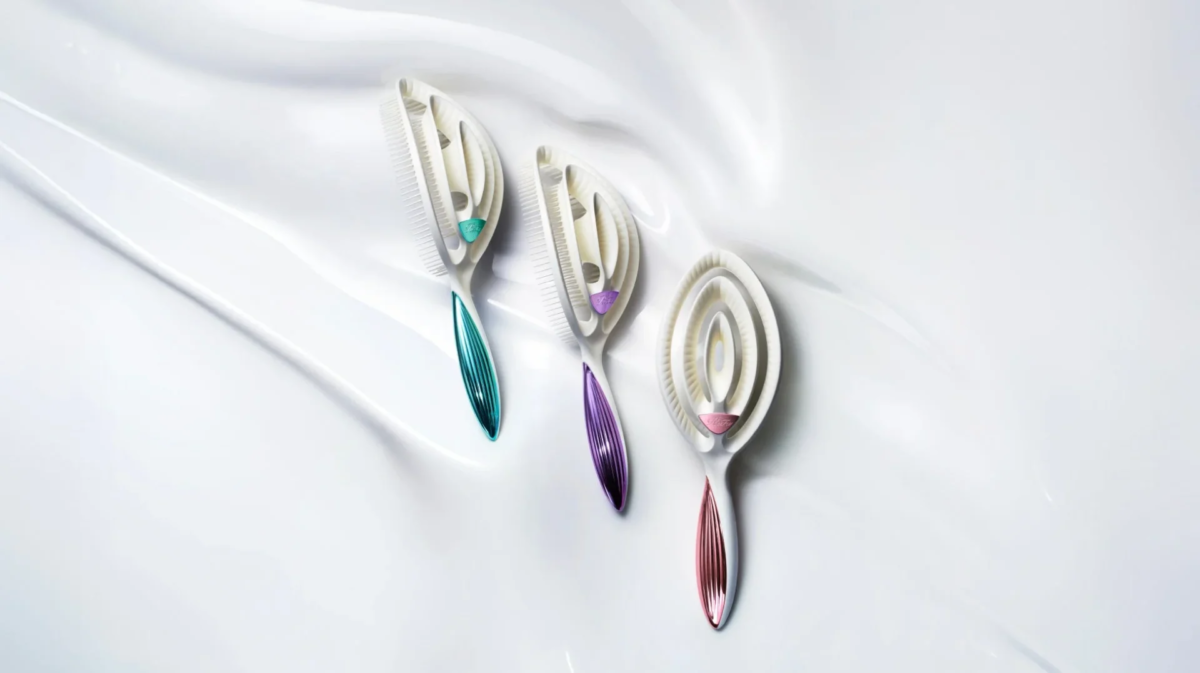 Tres cepillos de pelo de diseño moderno con mangos metálicos en verde, morado y rosa sobre una superficie blanca y ondulada.