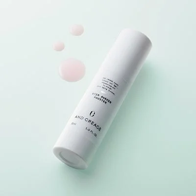 Botella blanca de And Creage Stem Awaken Booster con gotas de líquido rosado, sobre un fondo verde menta.