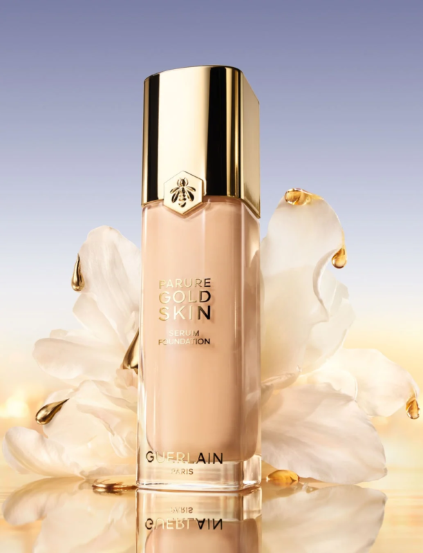 Botella de la base de maquillaje s&eacute;rum Parure Gold Skin de Guerlain con flores y gotas doradas